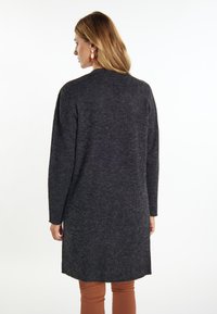 Cardigan lungo grigio scuro con maniche lunghe, materiale lavorato a maglia testurizzato e orlo curvo. Indossato sopra pantaloni aderenti arancio-marrone.