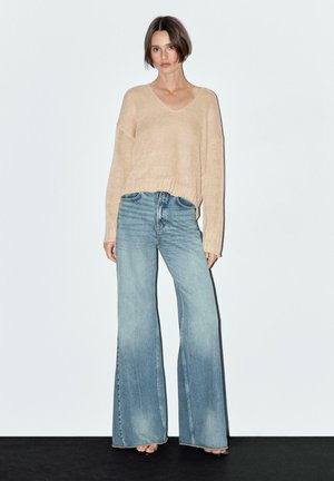 Beiger, gestrickter V-Ausschnitt-Pullover, kombiniert mit hellblauen, weiten Jeans. Der Pullover hat eine lockere Passform und gerippte Bündchen.
