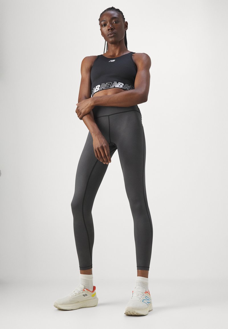 Soutien-gorge de sport noir avec logo sur bande blanche, associé à un legging noir ajusté taille haute. Des baskets blanches avec des accents colorés complètent le look.