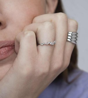 Anello d'argento caratterizzato da un design ondulato, adornato con piccole pietre preziose tonde e trasparenti. Mano poggiata sul viso, con una semplice fascia metallica su un altro dito.