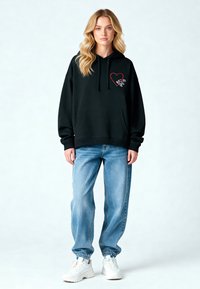 Felpa nera con cappuccio e tasca frontale, decorata con una grafica a forma di cuore rosso. Abbinata a jeans baggy azzurri chiari e sneaker bianche.