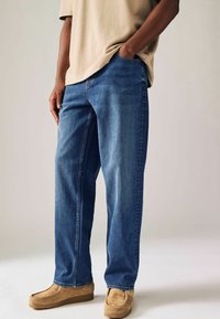Vaqueros de denim azul con corte recto, con un desvanecimiento sutil. Llevados con zapatos de ante beige sin cordones, combinados con una camiseta marrón claro.