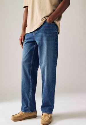 Jeans in denim blu con taglio dritto, caratterizzati da una leggera scoloritura. Indossati con scarpe slip-on in suede beige, abbinati a una T-shirt marrone chiaro.