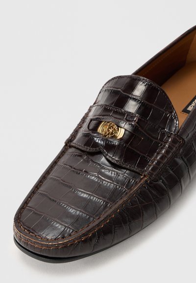 Mörkbrun loafers i läder med krokodilmönster, guldfärgad metallspänne och detaljerad sömnad på vit bakgrund.