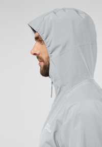 Lichtgrijze hooded jacket gemaakt van glad, waterafstotend materiaal. Voorzien van een koord bij de capuchon en een logo op de borst.
