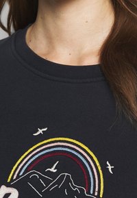 Tmavě námořnický svetr s vyšitým motivem hory a vícebarevným designem duhy. Doplněný bílými ptáky letícími nad ním.