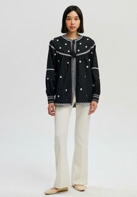 Black embroidered blouse with white floral patterns, round neck, and tassel tie, paired with fitted white pants and cream shoes.