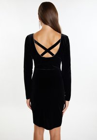 Schwarzes Samtkleid mit langen �Ärmeln, tiefem Rückenausschnitt, Kreuzträgern und einer taillierten Silhouette mit einer geschwungenen Saumlinie.