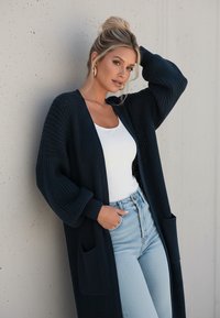 Granatowy kardigan dziergany z długimi rękawami, otwartym przodem, dwoma przednimi kieszeniami i strukturalną tkaniną, zestawiony z białą bluzką i jasnoniebieskimi dżinsami.