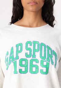 Felpa grigia chiara con la scritta "GAP SPORT 1969" in grande e vivace stampa verde e blu. Tessuto morbido con un classico design a collo rotondo.