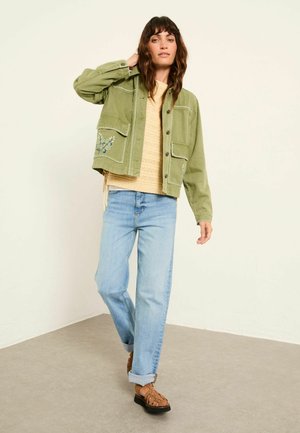 Green cropped jacket with embroidered details, paired with a textured beige sweater, light blue jeans, and brown woven sandals.