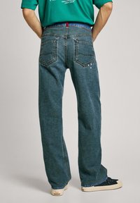 Jeans de mezclilla azul oscuro con un corte relajado, que cuentan con bolsillos traseros y sutiles acentos decorativos, combinados con zapatillas negras casuales.