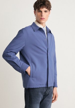 Blue lightweight jacket with a collar, snap buttons, and side pockets, paired with a light-colored shirt and tie. Smooth texture, casual design.