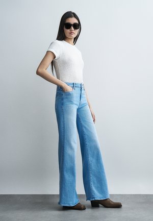 Γυναίκα που φοράει μαύρα γυαλιά ηλίου, λευκό κοντομάνικο τοπ, ανοιχτό μπλε τζιν με φαρδιά πόδια και καφέ μποτάκια με τακούνι, στέκεται με τα χέρια στις τσέπες.