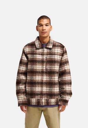 Jeune homme portant une veste zippée à carreaux marron et crème avec un col, par-dessus une chemise violette et un pantalon beige, sur un fond uni.