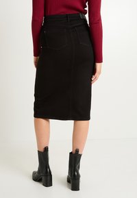 Falda lápiz de mezclilla negra con bolsillos traseros, cintura de altura media y costuras visibles. Combinada con una blusa de canalé color burdeos y botines negros de tacón.