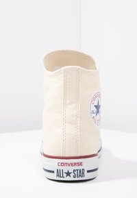 Baskets en toile beige avec un talon arrondi, présentant un patch logo circulaire, des accents rouges et bleus, et une semelle en caoutchouc blanche.