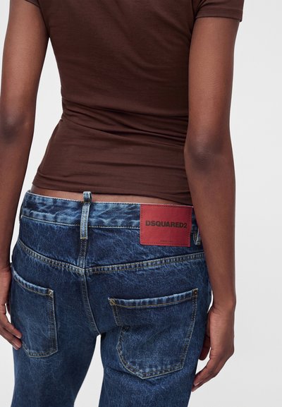 Personne portant un jean denim bleu avec une étiquette rouge DSQUARED2 sur la partie arrière droite de la ceinture et une chemise marron ajustée à manches courtes.