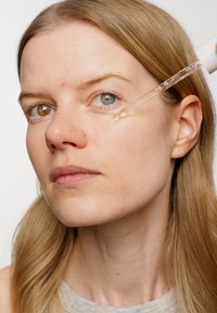Tydlig pipett som applicerar ett serum under en models öga. Modellen har rakt blont hår och varierande ögonfärger: bruna och blå. Slät hudstruktur.