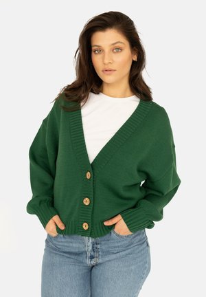 Zielony kardigan dzianinowy z głębokim dekoltem V, prążkowanymi akcentami i drewnianymi guzikami, noszony na białej koszuli, w zestawie z niebieskimi dżinsami.