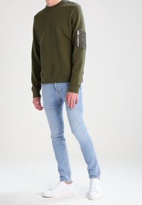Felpa verde oliva con scollo rotondo e tasca con zip sulla manica sinistra, abbinata a jeans azzurri e sneakers bianche.