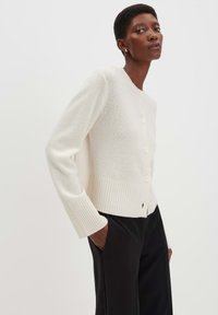 Frau in einem weißen, knöpfbaren Cardigan mit gerippten Bündchen und Saum, kombiniert mit schwarzen Hosen, steht mit den Händen in den Taschen.