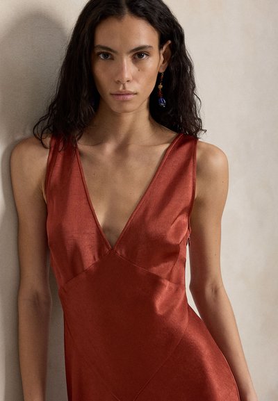 Γυναίκα με μακριά σκούρα μαλλιά, που φοράει αμάνικο σατέν φόρεμα σε σκουριά απόχρωση και κρεμαστά σκουλαρίκια, όρθια μπροστά σε μπεζ τοίχο.