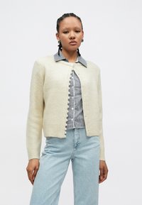 Kremen pleten pulover z okrolim izrezom, teksturirano tkanino in gumbi, ki spominjajo na bisere, v sprednjem delu, nošen čez srajco s karo ovratnikom.