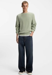 Suéter verde de punto con textura acanalada, escote redondo y mangas largas. Combinado con jeans oscuros de pierna ancha y zapatos casuales.