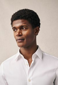 Uomo con capelli neri corti e ricci, che indossa una camicia bianca con bottoni, guarda di lato su uno sfondo chiaro e semplice.
