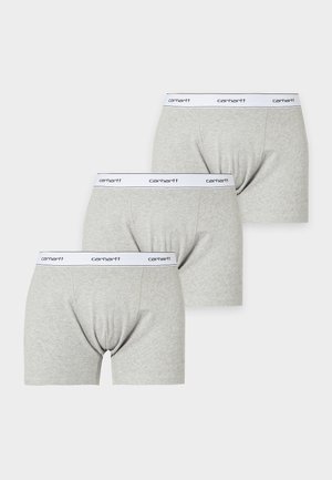 Homme mince à la peau foncée, debout pieds nus, portant un boxer gris clair avec une ceinture blanche dans un studio blanc simple.