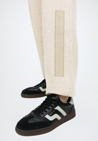 Ασπρόμαυρα sneakers GANT φορεμένα με μπεζ παντελόνι που έχουν κάθετο ορθογώνιο λογότυπο στο κάτω μέρος του ποδιού.
