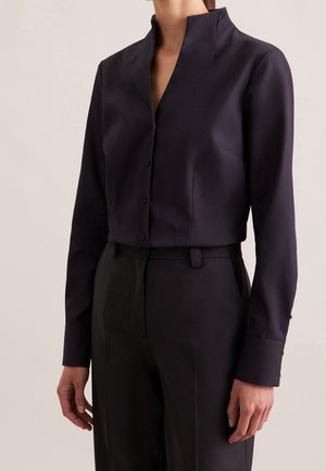 Vrouw draagt een nauwsluitend, donkerblauw, hemd met lange mouwen, een hoge kraag en op maat gemaakte zwarte broek.