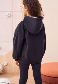 Sweat à capuche noir à coupe décontractée avec poignets côtelés, associé à des leggings noirs. L'arrière-plan présente un bois clair et des accessoires circulaires.