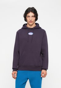 Jovem vestido com um hoodie roxo escuro com um pequeno logótipo multicolorido e calças azuis brilhantes, de pé contra um fundo branco simples.