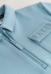 Hellblauer Pullover mit strukturiertem Stoff, ausgestattet mit einem frontalen Reißverschluss und einem rechteckigen Logo-Patch mit der Aufschrift "BAKER." Kragen und Nähte sind sichtbar.