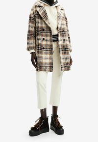 Cappotto doppiopetto a quadri in crema, arancione e nero, indossato sopra un maglione crema con un motivo a trecce e orlo nero. Pantaloni bianchi e stivali alla caviglia neri con decorazioni completano il look.