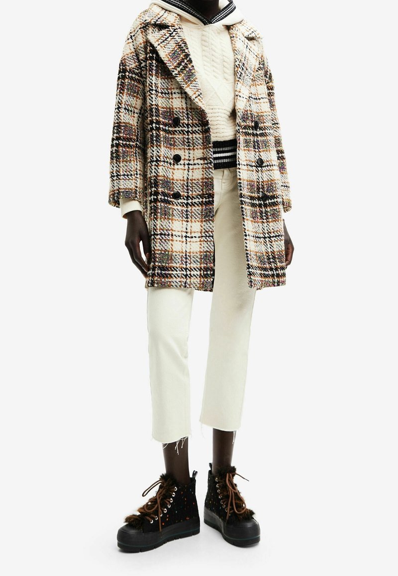 Cappotto doppiopetto a quadri in crema, arancione e nero, indossato sopra un maglione crema con un motivo a trecce e orlo nero. Pantaloni bianchi e stivali alla caviglia neri con decorazioni completano il look.