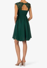 Grön klänning med spetsärmar och smockad liv. Flödande kjol med en utskärning bak. Buren med klara klacksandaler.