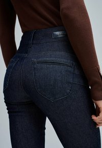 Jeans de mezclilla azul oscuro con una cintura alta, con costuras detalladas y dos bolsillos traseros. Etiqueta negra sujeta en la cinturilla.