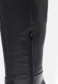 Botte noire en cuir jusqu'au genou avec fermeture éclair sur le côté, texture lisse et design épuré avec un subtil détail de couture sur le tige.
