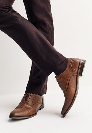 Brune læder brogue sko båret med mørkebrune skræddersyede bukser, den ene fod fladt på gulvet og den anden hævet på hælen.