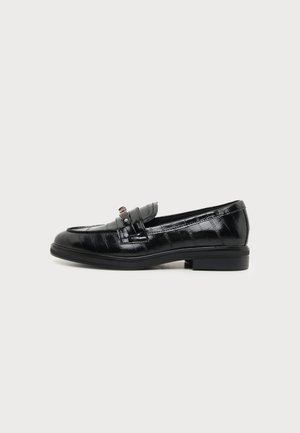 Mocasín de cuero negro con tacón bajo y una pieza metálica decorativa en la parte superior, mostrado de perfil sobre un fondo liso.