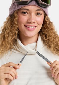 Jasnobeżowa bluza z kapturem z wysokim kołnierzem, wyposażona w sznurek ze wzorem jodełki w czarno-białych kolorach. Modelka z kręconymi włosami nosząca fioletową czapkę beanie.