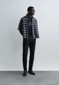 Άνδρας που φοράει μαύρα γυαλιά ηλίου, ασπρόμαυρο καρό πουκάμισο πάνω από μαύρο μπλουζάκι με στάμπα, μαύρο παντελόνι και μαύρα μποτάκια, στέκεται μπροστά σε απλό τοίχο.