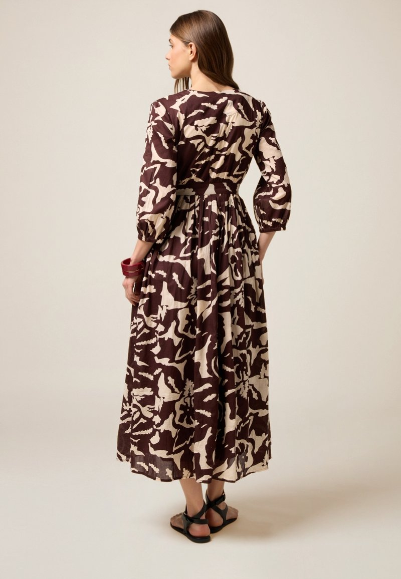 Vestido maxi en estampado floral marrón y crema, confeccionado en una tela ligera, con mangas abullonadas, cintura ajustada con cinturón y un dobladillo fluido.