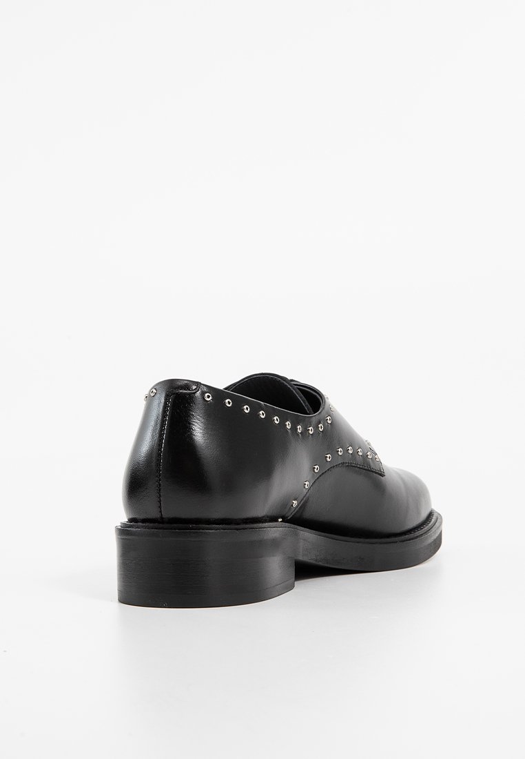 Chaussure en cuir noir avec bout rond, ornée de clous en argent le long du bord supérieur et à semelle épaisse en caoutchouc. Texture lisse dans l'ensemble.
