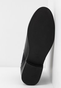 Semelle de chaussure noire avec un talon texturé et un avant lisse, photographiée sur un fond blanc en coin.