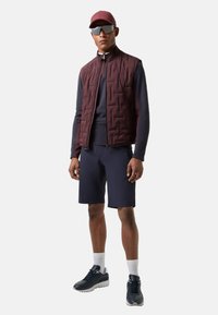 Gesteppte bordeauxrote Weste über einem marineblauen Langarmshirt und passenden Shorts. Dunkle Sneakers und eine Baseballkappe mit Sonnenbrille vervollständigen den Look.
