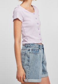 Světle fialový žebrovaný crop top s krátkými rukávy a třepeným lemem, ozdobený pěti odpovídajícími knoflíky. Slučuje se s světle modrými džínovými šortkami s vysokým pasem.